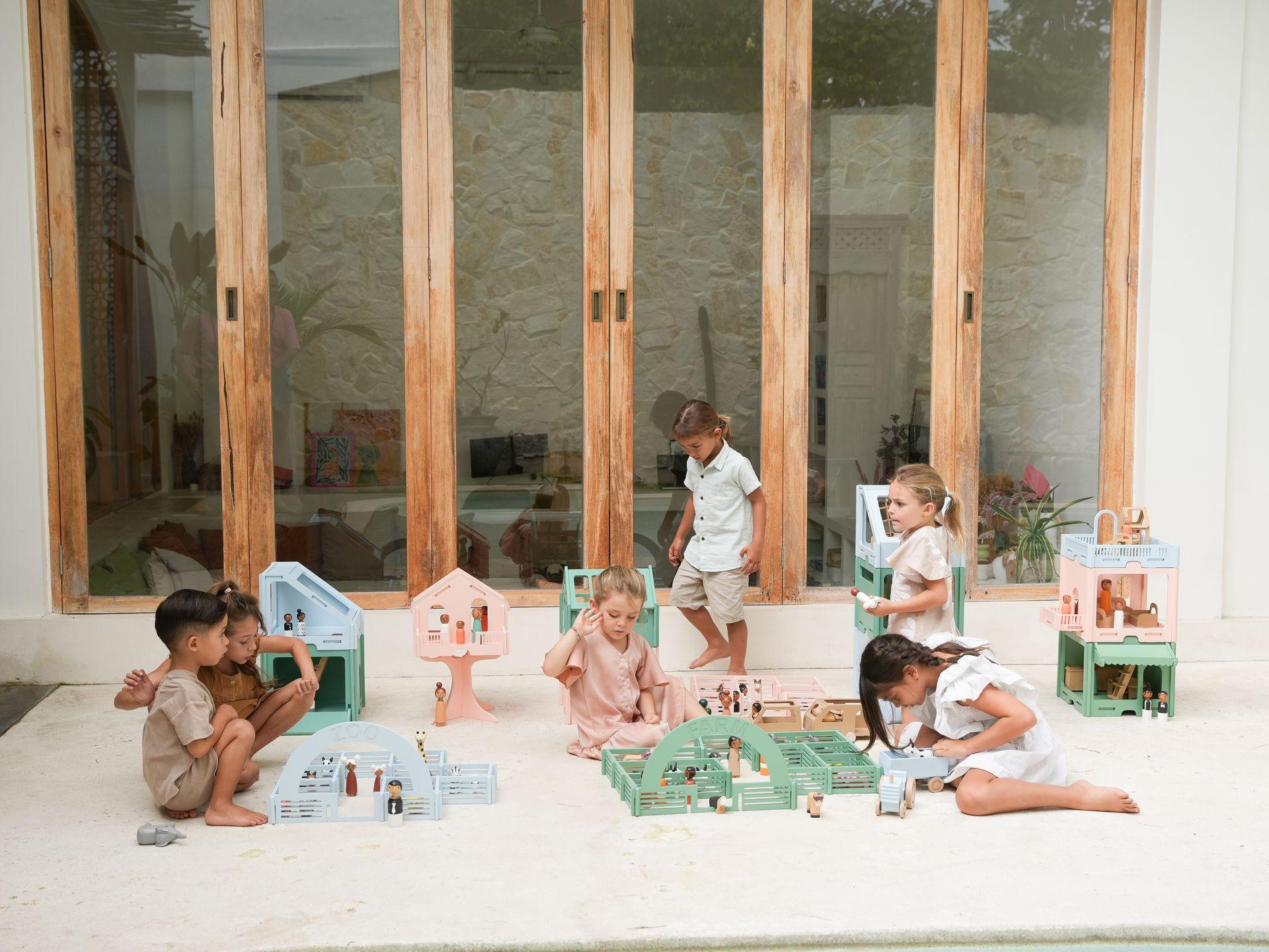 Zes kinderen spelen met houten speelgoedsetjes in een lichte buitenruimte met grote glazen deuren op de achtergrond. De kinderen, variërend in leeftijd en gender, zitten of hurken op de grond en zijn verdiept in hun spel. Het speelgoed bestaat uit miniatuurhuisjes, boerderijen en figuren, voornamelijk in pastelkleuren zoals lichtblauw, roze en mintgroen. De omgeving is minimalistisch met een stenen muur door de glazen deuren en een lichte tegelvloer.
