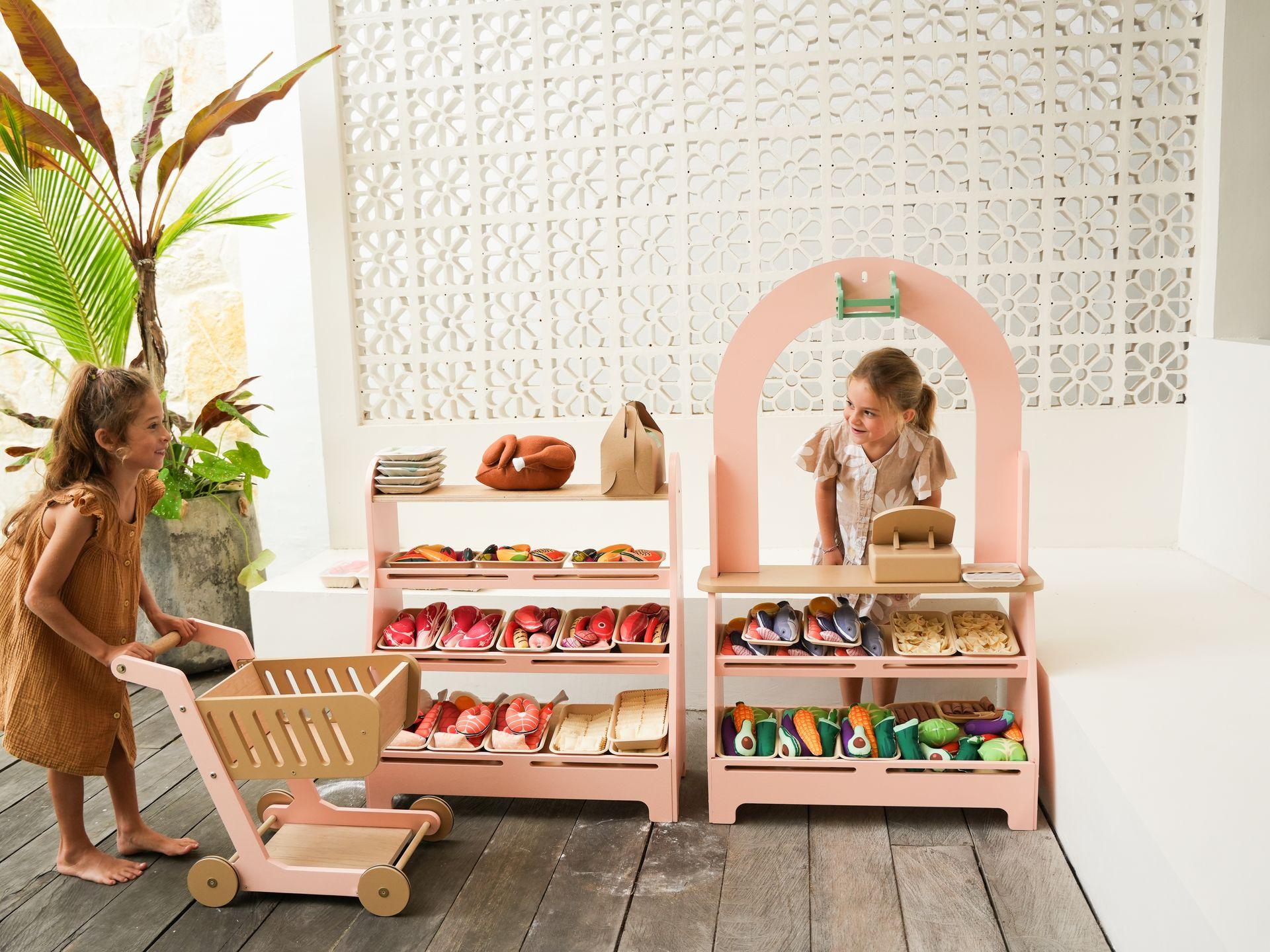 Twee jonge meisjes spelen in een openluchtwinkel. Het meisje links duwt een houten winkelwagentje en lacht. Het meisje rechts staat achter een roze houten marktkraam met twee planken vol met stoffen speelgoedeten. De marktkraam heeft een gebogen roze frame. De hele scène speelt zich af in een lichte, open ruimte met planten en een witte, gedetailleerde muur op de achtergrond.