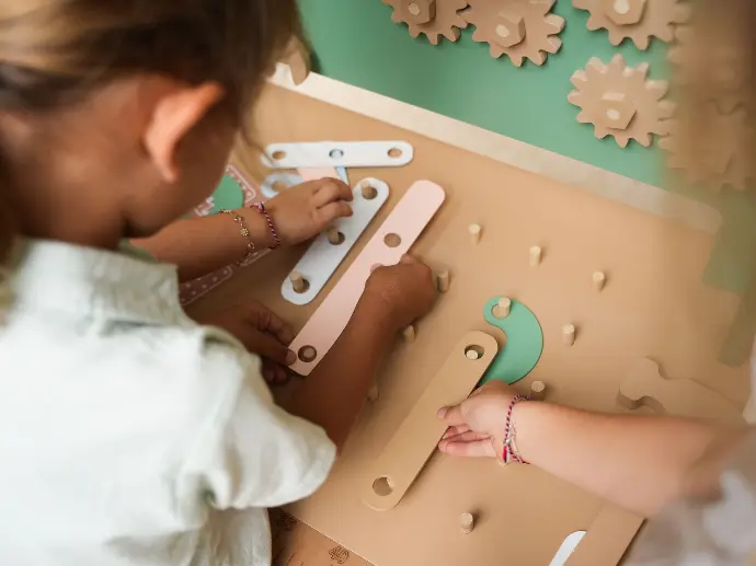 Close-up van kinderen die samenwerken aan een houten werkbank met pastelgekleurde gereedschappen en onderdelen, terwijl ze een bouwtekening volgen.