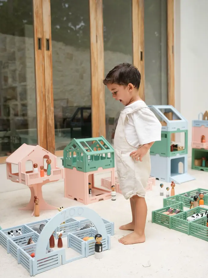 Een jongetje speelt enthousiast met pastelgekleurde houten speelsets, waaronder een dierentuin en poppenhuizen, in een lichte buitenomgeving.