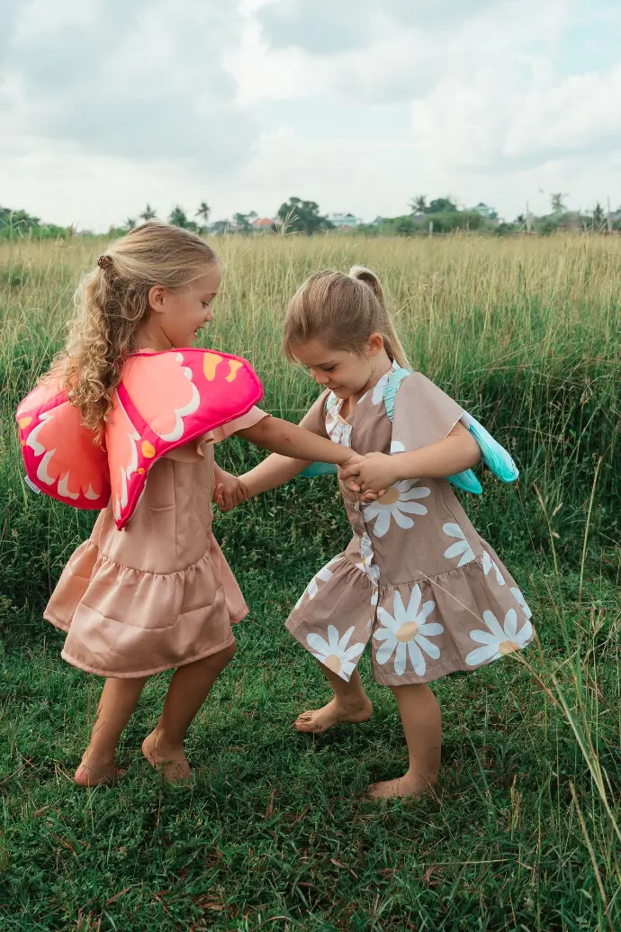 Twee jonge meisjes met vleugels op hun rug houden elkaars hand vast en lijken te dansen of te spelen in een hoog grasveld. Het meisje links heeft lang blond krullend haar en draagt een roze jurk met felroze en oranje vlindervleugels. Het meisje rechts heeft blond haar vastgebonden in een paardenstaart en draagt een bruine jurk met witte madeliefjespatronen en lichtblauwe vleugels. Beiden kijken naar elkaar en glimlachen. De achtergrond bestaat uit weelderig groen gras en een bewolkte lucht.
