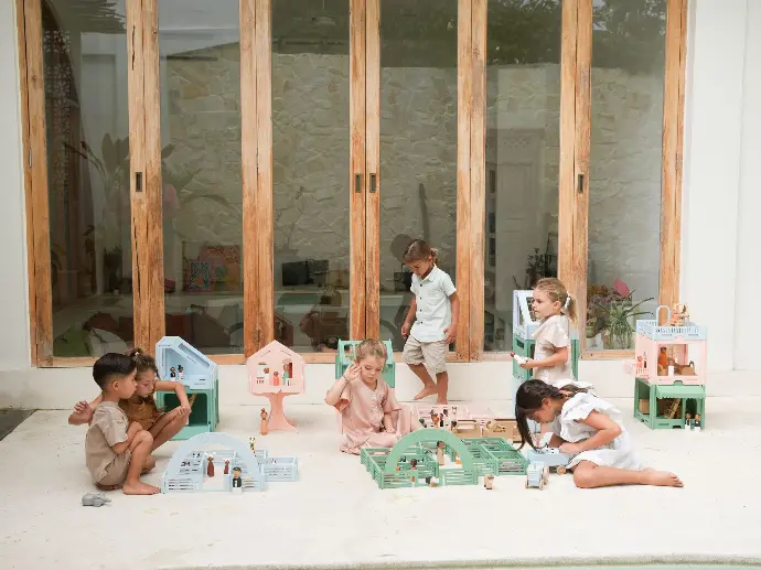 Zes kinderen spelen met houten speelgoedsetjes in een lichte buitenruimte met grote glazen deuren op de achtergrond. De kinderen, variërend in leeftijd en gender, zitten of hurken op de grond en zijn verdiept in hun spel. Het speelgoed bestaat uit miniatuurhuisjes, boerderijen en figuren, voornamelijk in pastelkleuren zoals lichtblauw, roze en mintgroen. De omgeving is minimalistisch met een stenen muur door de glazen deuren en een lichte tegelvloer.